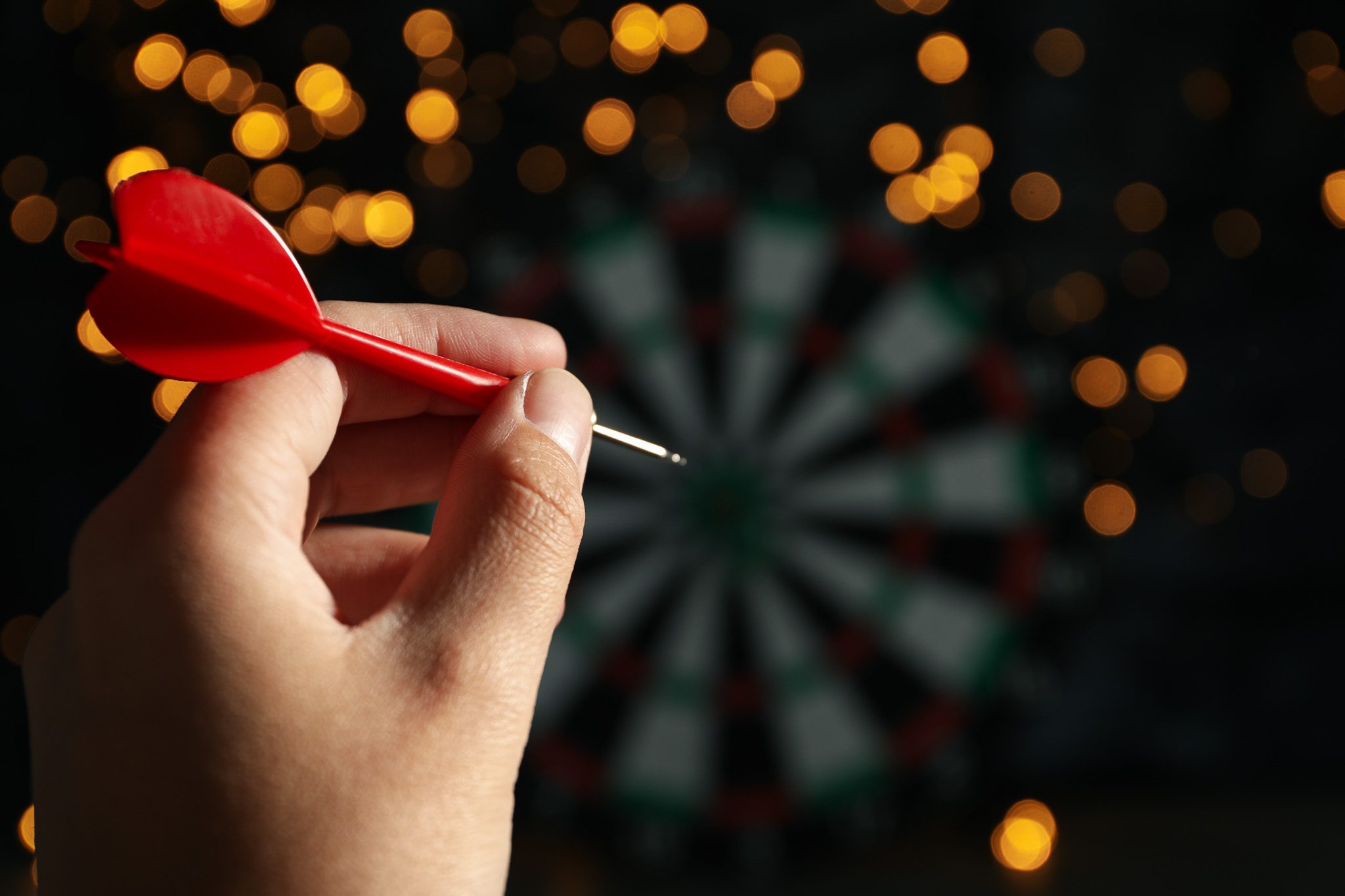 A hand holding a red dart aimed at a dartboard, with a backdrop of blurred warm golden lights creating a bokeh effect, evoking the relaxed atmosphere of an Auckland pub.